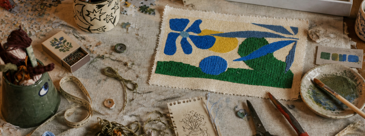 Imagem mostra uma mesa de trabalho artesanal com um bordado colorido de formas abstratas em azul, verde e amarelo ao centro, cercado por linhas, botões, tesoura, desenhos e materiais de craft, destacando o processo manual e criativo.